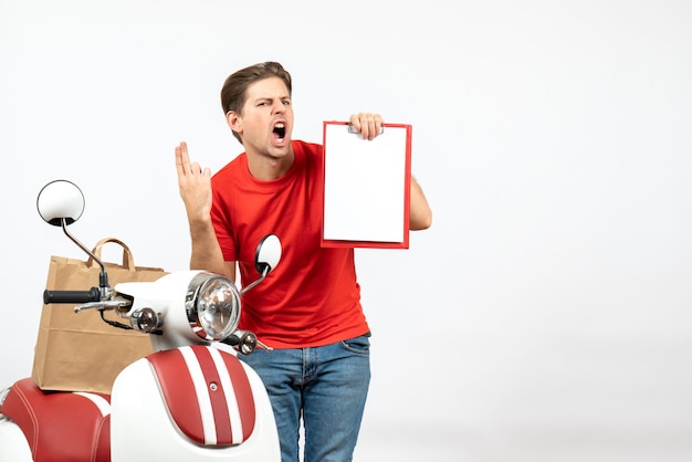 Repartidor nervioso joven en uniforme rojo de pie cerca de scooter sosteniendo el documento que muestra dos en la pared blanca