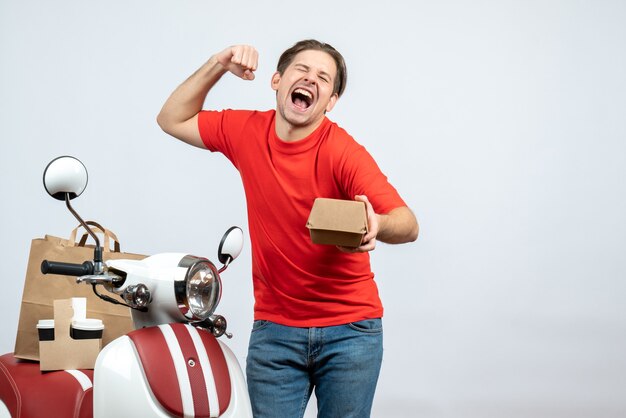 Repartidor orgulloso en uniforme rojo de pie cerca de scooter sosteniendo una pequeña caja sobre fondo blanco.