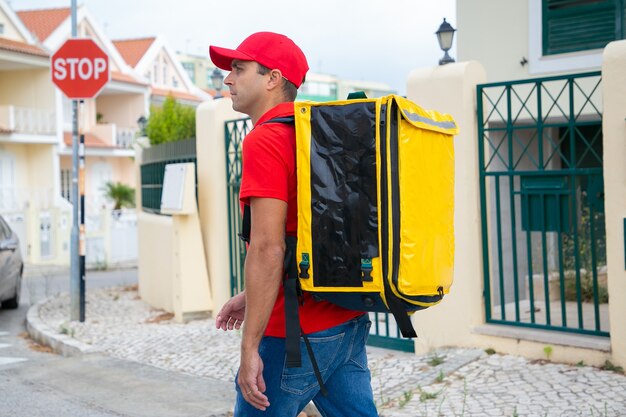 Repartidor pensativo con gorra roja mirando la casa requerida. Mensajero de mediana edad con mochila térmica amarilla entregando pedidos urgentes y caminando por la calle. Servicio de entrega y concepto de compra online.