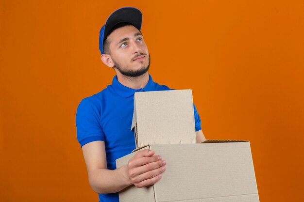 Repartidor pensativo joven vistiendo polo azul y gorra de pie con cajas mirando hacia arriba sobre fondo naranja aislado