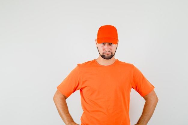 Foto gratuita repartidor de pie con las manos en la cintura en camiseta naranja, gorra y mirando nervioso. vista frontal.