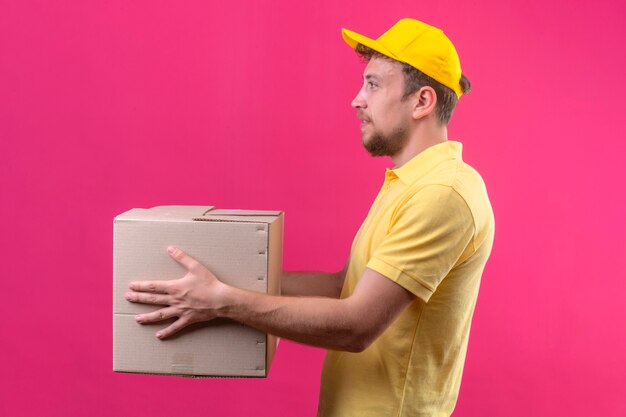 Repartidor en polo amarillo y gorra dando caja de cartón grande a un cliente de pie de lado en rosa