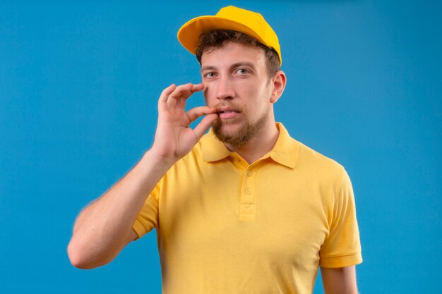 Repartidor en polo amarillo y gorra haciendo gesto de silencio haciendo como cerrar la boca con una cremallera en azul aislado