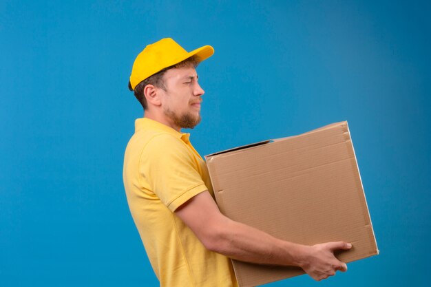 Repartidor en polo amarillo y gorra de pie con gran caja de cartón que sufren de gran peso en azul