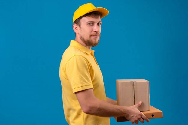 Repartidor en polo amarillo y gorra de pie lateralmente con cajas de cartón sonriendo amable en azul