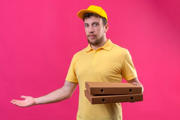 Repartidor en polo amarillo y gorra sosteniendo cajas de pizza y presentando con la mano y la palma de pie en rosa