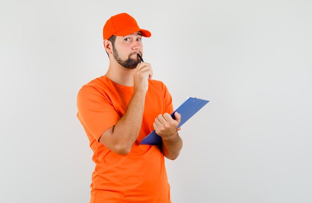 Repartidor con portapapeles y bolígrafo en camiseta naranja, gorra y mirando pensativo. vista frontal.