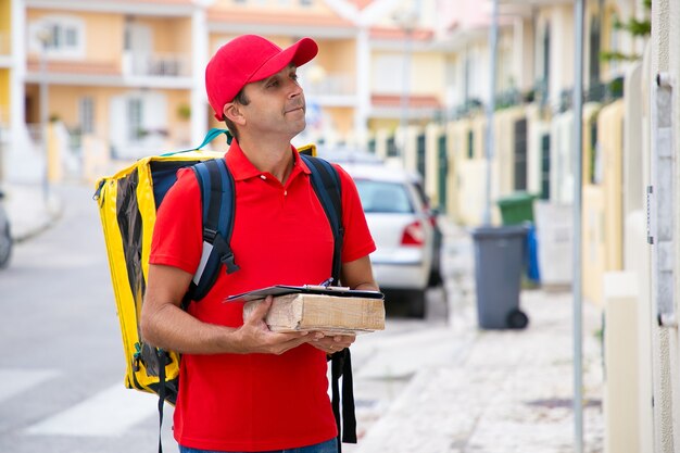 Repartidor positivo sosteniendo el paquete con el portapapeles y esperando al destinatario al aire libre. Hombre caucásico guapo con mochila amarilla entregando pedidos a la gente. Servicio de entrega y concepto de correo.