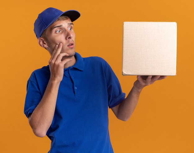 Repartidor rubio joven ansioso pone la mano en la cara sosteniendo y mirando la caja de cartón aislada en la pared naranja con espacio de copia