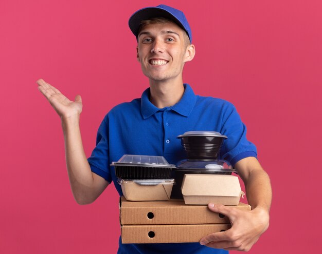 Repartidor rubio joven sonriente está parado con la mano levantada y sostiene los envases y paquetes de comida en cajas de pizza