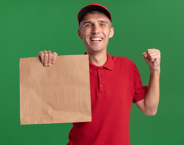 Repartidor rubio joven sonriente mantiene el puño y sostiene el paquete de papel en verde