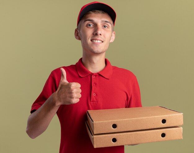 Repartidor rubio joven sonriente pulgar hacia arriba y sostiene cajas de pizza en verde oliva