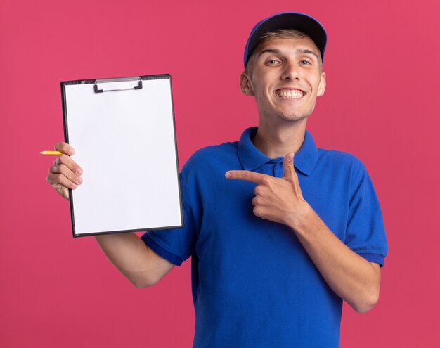 Repartidor rubio joven sonriente sostiene y apunta al portapapeles