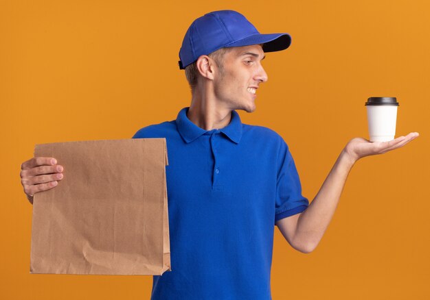 Repartidor rubio joven sonriente sostiene el paquete de papel y mira la taza en naranja