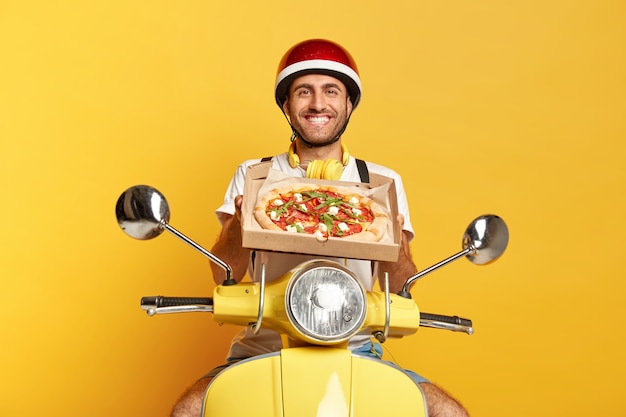 Repartidor satisfecho con casco conduciendo scooter amarillo mientras sostiene la caja de pizza