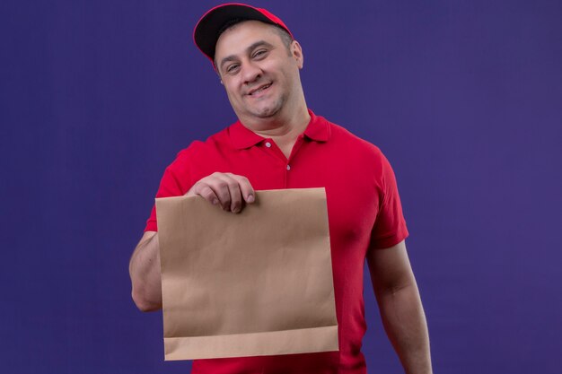 Repartidor satisfecho vistiendo uniforme rojo y gorra con paquete de papel positivo y feliz sonriendo