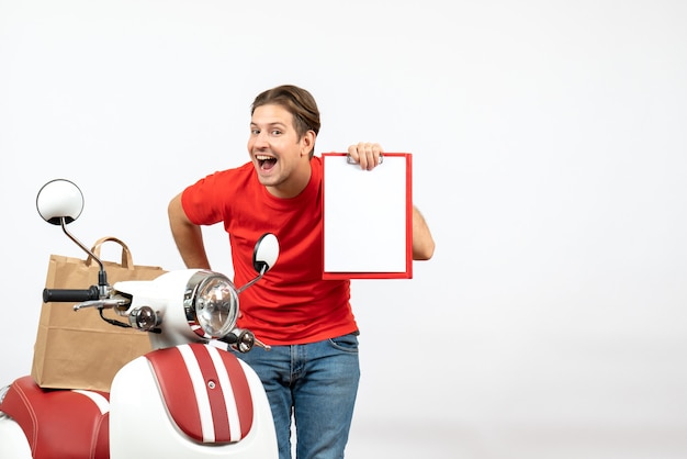 Repartidor sonriente confiado joven en uniforme rojo que se coloca cerca del scooter que muestra el documento en la pared blanca