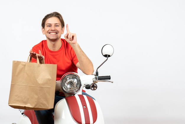 Repartidor sonriente joven en uniforme rojo sentado en scooter sosteniendo una bolsa de papel y apareciendo en la pared blanca