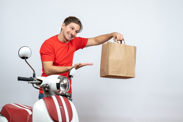 Repartidor sonriente en uniforme rojo de pie cerca de scooter señalando la caja de papel sobre fondo blanco.