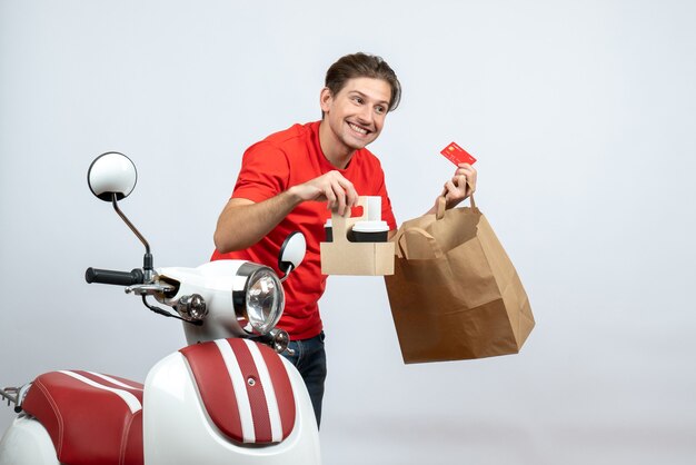 Repartidor sonriente en uniforme rojo de pie cerca de scooter y sosteniendo pedidos con tarjeta bancaria sobre fondo blanco.