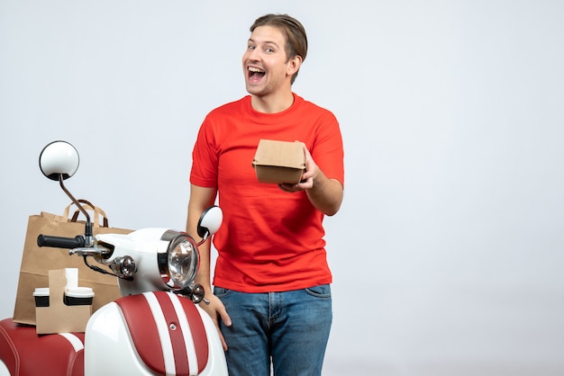Foto gratuita repartidor sonriente en uniforme rojo de pie cerca de scooter sosteniendo una pequeña caja sobre fondo blanco.