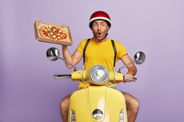 Repartidor sorprendido conduciendo scooter amarillo mientras sostiene la caja de pizza