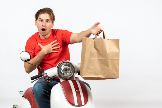 Repartidor sorprendido joven en uniforme rojo de pie cerca de scooter dando bolsa de papel a alguien en la pared blanca