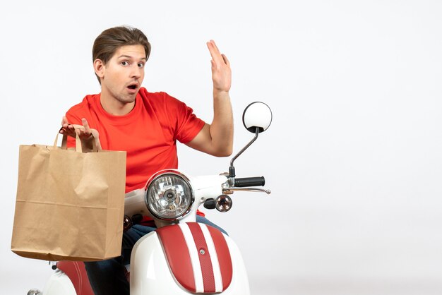 Repartidor sorprendido joven en uniforme rojo sentado en scooter sosteniendo una bolsa de papel y mostrando cinco en la pared blanca