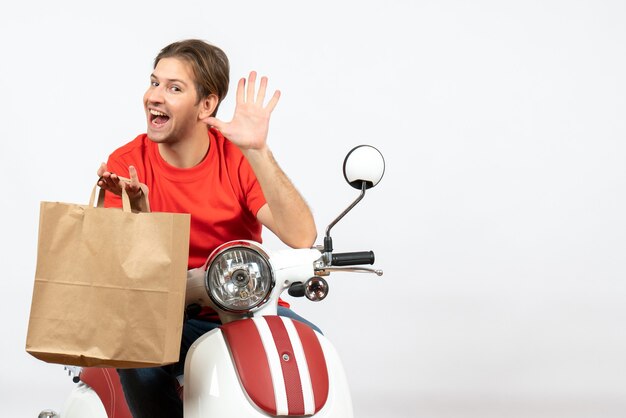 Repartidor sorprendido joven en uniforme rojo sentado en scooter sosteniendo una bolsa de papel y mostrando cinco en la pared blanca