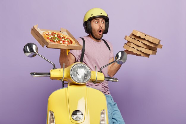 El repartidor sorprendido lleva una pila de deliciosa pizza italiana, usa casco y ropa casual, conduce una motocicleta, transporta comida rápida para la cena, aislado sobre una pared púrpura. Bocadillo sabroso