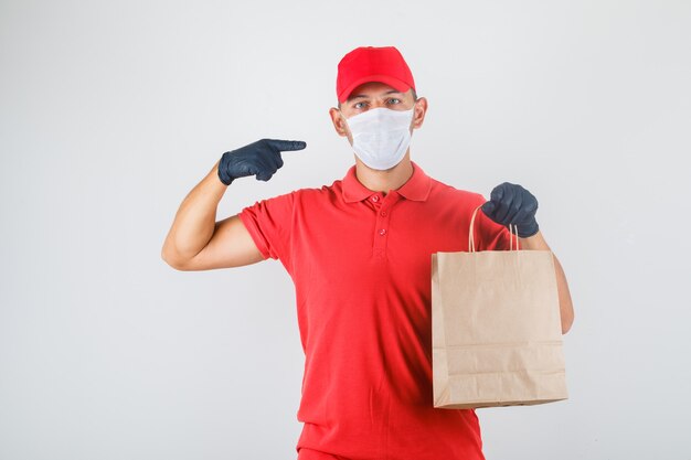 Repartidor sosteniendo una bolsa de papel y apuntando a sí mismo con uniforme rojo, máscara médica, guantes