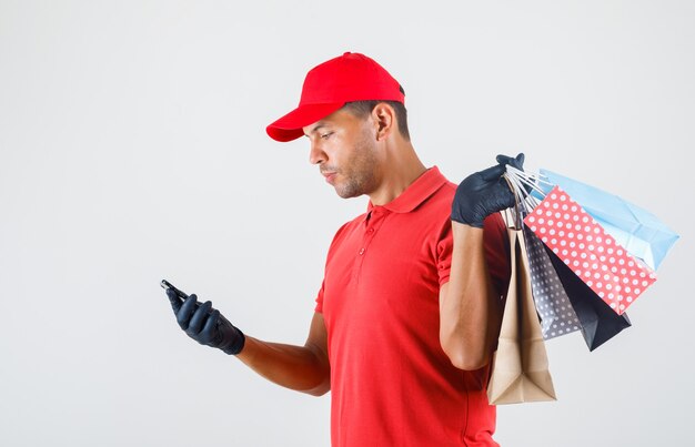 Repartidor sosteniendo bolsas de papel y mirando smartphone en uniforme rojo, guantes.
