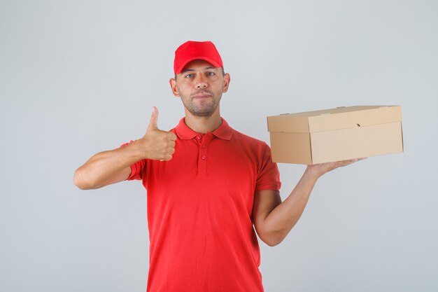 Repartidor sosteniendo una caja de cartón y mostrando el pulgar hacia arriba en uniforme rojo y mirando positivo.