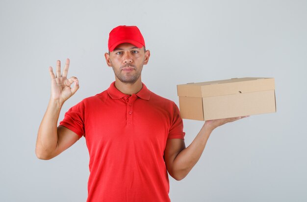 Repartidor sosteniendo una caja de cartón y mostrando el signo de ok en uniforme rojo