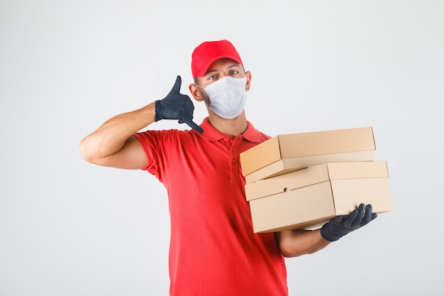 Repartidor sosteniendo cajas de cartón y haciendo señal de llamada en uniforme rojo, máscara médica, guantes vista frontal.