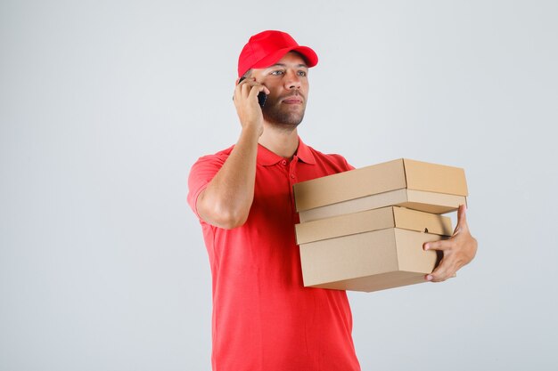 Repartidor sosteniendo cajas de cartón mientras habla por teléfono celular en uniforme rojo