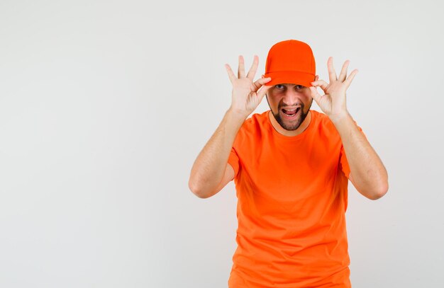 Repartidor sosteniendo los dedos en su gorra en camiseta naranja y mirando confiado, vista frontal.
