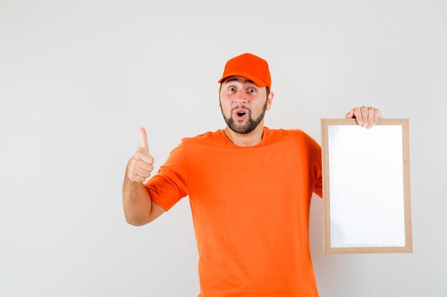 Repartidor sosteniendo el marco en blanco con el pulgar hacia arriba en camiseta naranja, gorra y mirando feliz, vista frontal.