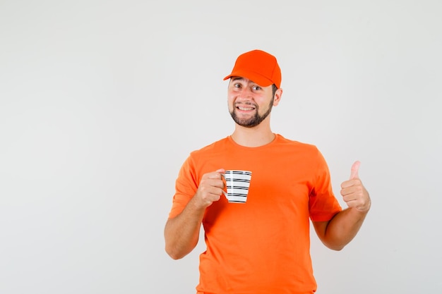 Repartidor sosteniendo una taza de bebida con el pulgar hacia arriba en camiseta naranja, gorra y mirando alegre, vista frontal.