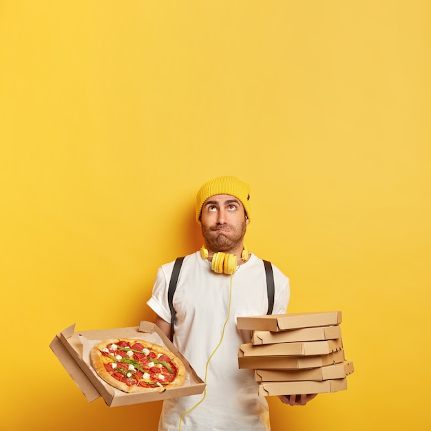 El repartidor trae cajas de cartón para pizza para el cliente, mira hacia arriba, usa sombrero amarillo, camiseta blanca, trabaja transportando comida rápida, aislado en la pared amarilla, copia espacio para su promoción