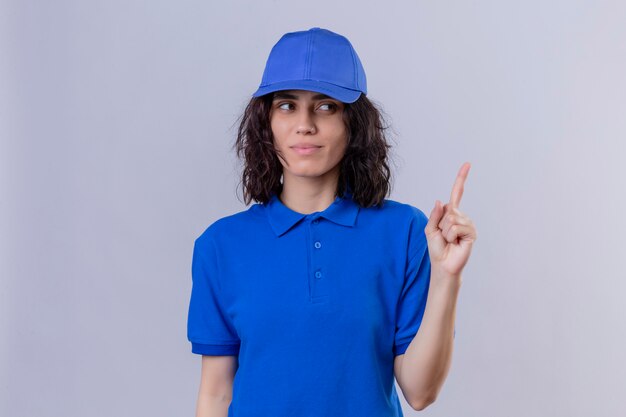 Repartidor en uniforme azul y gorra apuntando hacia arriba con el dedo sonriendo confiado concentrado en la tarea de pie en blanco