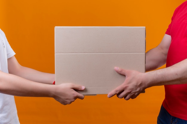 Foto gratuita repartidor en uniforme rojo dando paquete caja a un cliente sobre pared naranja aislada