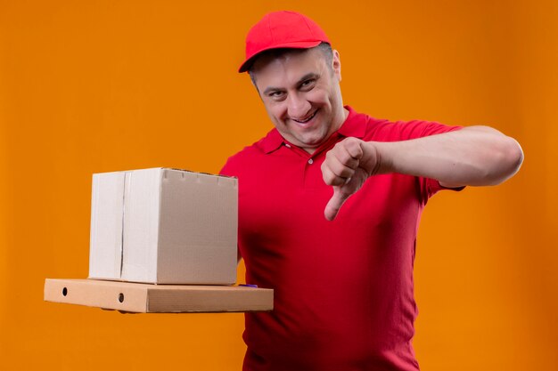 Repartidor con uniforme rojo y gorra con caja de cartón y cajas de pizza sonriendo mostrando los pulgares hacia abajo mirando astutamente a la cámara sobre la pared naranja