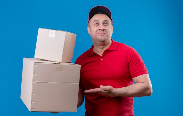 Repartidor con uniforme rojo y gorra con cajas de cartón apuntando hacia él con el brazo de su mano sonriendo mirando confundido sobre la pared azul
