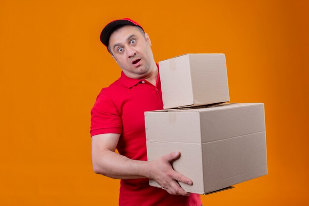 Repartidor con uniforme rojo y gorra con cajas de cartón conmocionados con los ojos bien abiertos sobre la pared naranja aislada