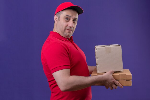 Repartidor con uniforme rojo y gorra con cajas de cartón sorprendido sobre pared púrpura aislado