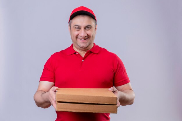 Repartidor con uniforme rojo y gorra con cajas de pizza sonriendo amable con cara feliz sobre pared blanca aislada