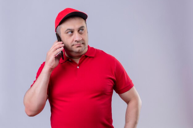 Repartidor en uniforme rojo y gorra hablando por teléfono móvil mirando a un lado con cara seria sobre pared blanca