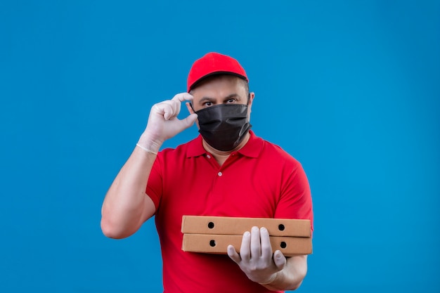 Repartidor con uniforme rojo y gorra en máscara protectora facial con cajas de pizza gesticulando con la mano mostrando un signo de tamaño pequeño, símbolo de medida sobre la pared azul