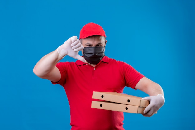 Repartidor con uniforme rojo y gorra en máscara protectora facial con cajas de pizza mostrando los pulgares hacia abajo sobre la pared azul